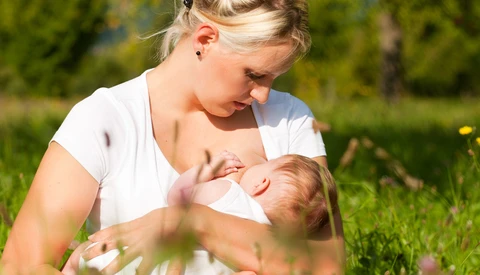 Mother breastfeeding infant outdoors in natural daylight surrounded by grass and greenery.