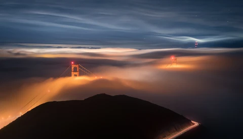 Golden Gate Bridge emerging from thick fog at twilight, illuminated by warm sunset light above a dark hillside silhouette.