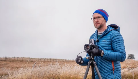 Filmmaker in blue jacket and beanie operating a cinema camera on tripod in an open field.