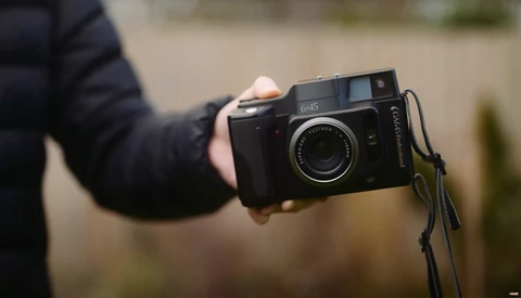 Photographer's hand holding a black rangefinder camera with a lens cap on top and shoulder strap attached.