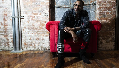 Man in dark patterned shirt seated on red Chesterfield armchair against exposed brick wall.