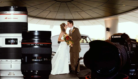 Wedding couple embracing under a modern dome structure, photographed through an array of Canon lenses and camera equipment in the foreground.