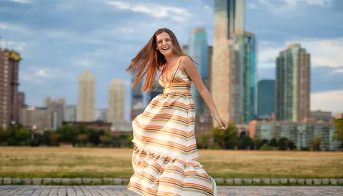 Woman in striped sundress poses on waterfront promenade with urban skyline behind her.