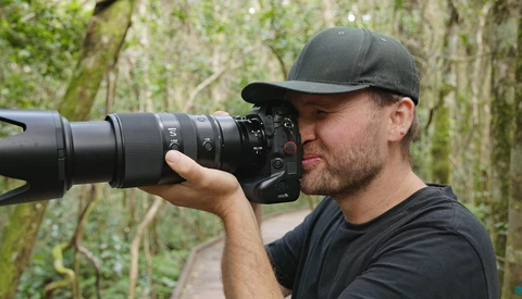 Photographer holding a DSLR camera with telephoto lens up to his eye while shooting outdoors.