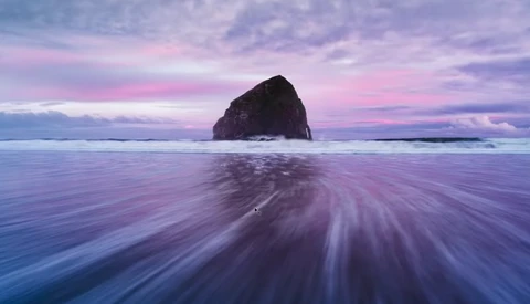 A dark rocky sea stack rises from the ocean during twilight, with smooth water motion blur in the foreground.