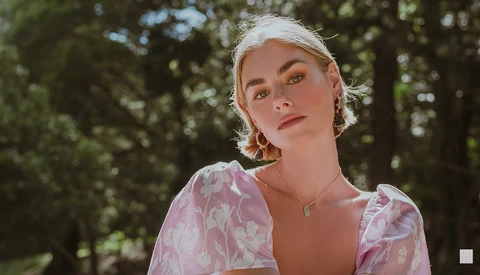 Portrait of a woman with platinum blonde hair wearing a pink patterned blouse against a blurred forest backdrop.