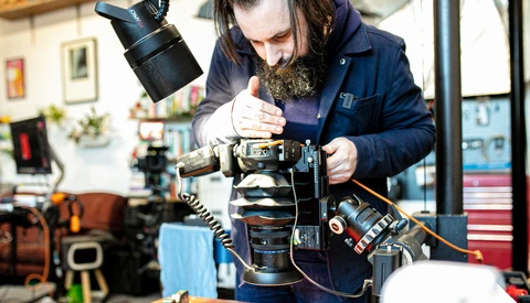 Photographer wearing navy jacket examines camera lens components at a workbench in a studio setting.