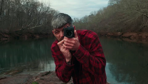 Photographer in red plaid shirt crouching by a river, aiming a telephoto lens toward the camera.