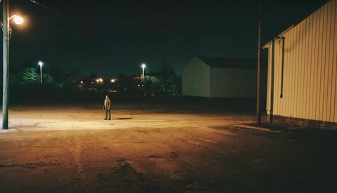 Solitary figure standing in an empty lit parking area at night beside an industrial metal building.