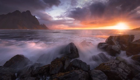 Rocky coastline at sunset with waves washing over dark boulders and dramatic cloudy sky.