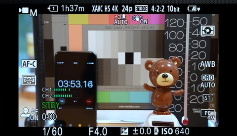 Camera LCD screen displaying a brown teddy bear figurine during video recording with audio levels and exposure settings visible.