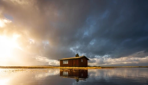 Solitary red-brown cabin reflected in still water under dramatic cloudy sky with golden sunlight breaking through.