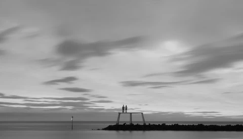 Two silhouetted figures stand on a concrete structure extending into calm water, dramatic clouded sky above.
