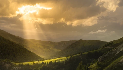 Mountain valley with golden sunlight breaking through clouds, forested slopes, and meadow in afternoon light.