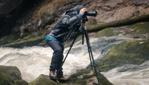 Photographer on tripod shooting whitewater rapids flowing over moss-covered rocks.