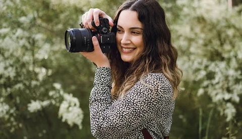 Woman holding a professional camera with telephoto lens, smiling at camera against blooming white flowers.