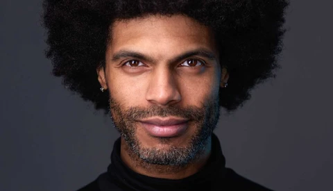 Portrait of a man with natural afro hair and facial hair against a neutral gray background.