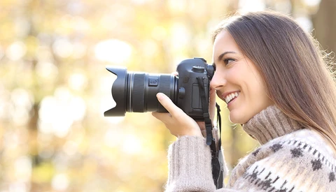 Woman holding a DSLR camera to her face while smiling outdoors.
