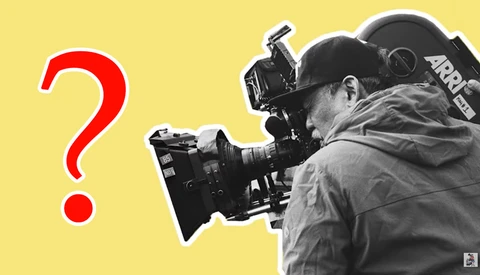 Cinematographer operating a professional film camera while wearing winter clothing against a yellow background.
