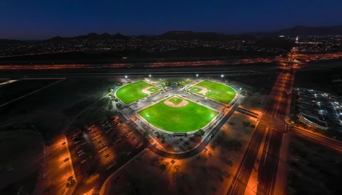 Aerial nighttime view of illuminated sports complex with three rectangular fields surrounded by parking areas and city lights.