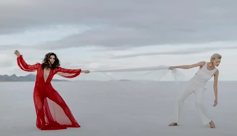 Two dancers in flowing clothing pose on a minimalist salt flat with arms extended in opposite directions.