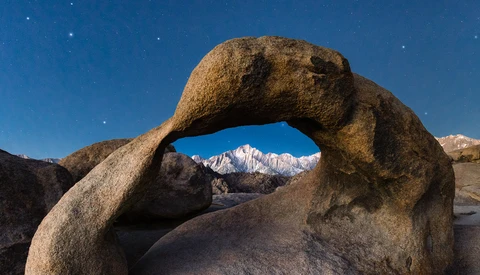 Granite boulder arch frames snow-capped mountain peak under starry night sky.