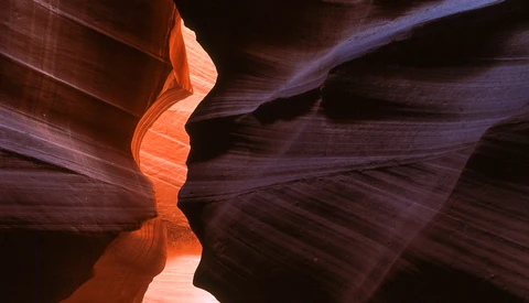 Slot canyon with undulating sandstone walls lit by warm orange light filtering from above, creating dramatic shadows and deep purple tones.