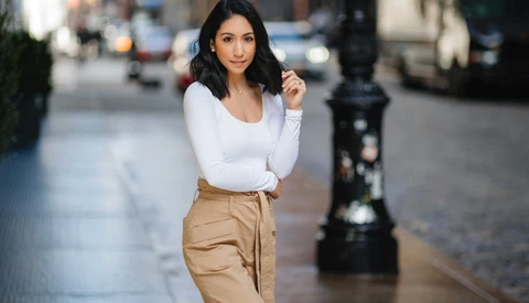 Woman in white long-sleeve shirt and tan pants standing on an urban street, smiling at camera.