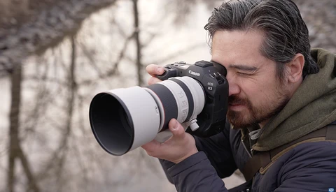 Photographer holding a DSLR camera with a white telephoto lens, composing a shot outdoors.