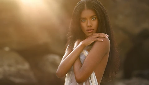 Woman with long dark hair wearing a white sleeveless top, posed against a warm, backlit background.