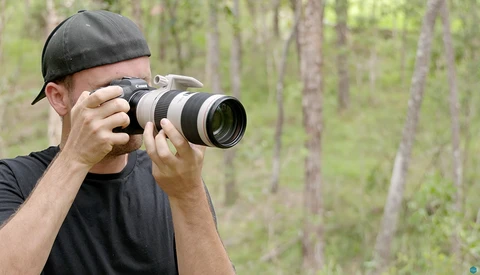 Photographer holding a telephoto lens camera to his face in a forest setting.