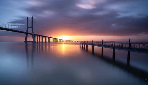 Modern cable-stayed bridge at sunrise with still water reflecting golden light and dramatic clouds.