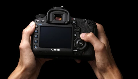Photographer's hands holding a Canon DSLR camera from behind, displaying the rear LCD screen and controls against a black background.