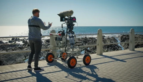 Filmmaker gestures while operating a camera rig mounted on a wheeled dolly at a coastal location.