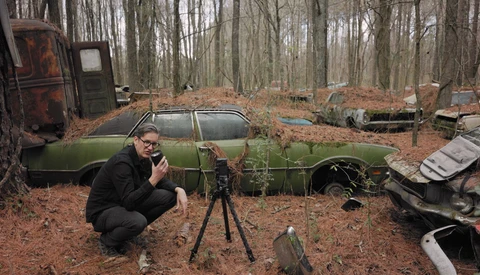 Photographer crouching beside a tripod-mounted camera in an abandoned car graveyard surrounded by bare trees.
