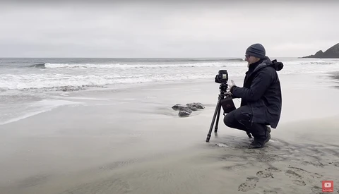 Photographer crouching on a beach with a tripod-mounted camera while framing ocean waves.