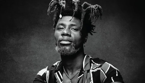 Black and white portrait of a man with styled dreadlocks, beard, and patterned shirt against dark background.