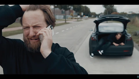 Man in dark clothing on phone with car and person visible in background on roadside.