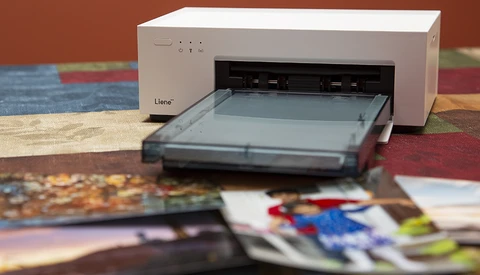 Compact photo printer with paper tray open, positioned over scattered printed photographs and colorful background.