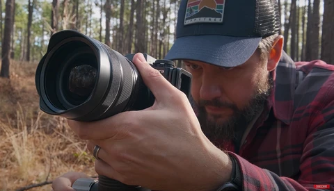 Photographer holding a mirrorless camera with telephoto lens, framing a shot in a forested woodland setting.