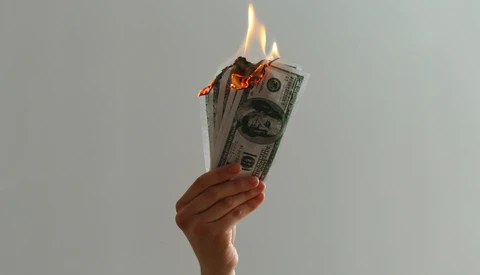 Hand holding burning U.S. dollar bills against a neutral background.