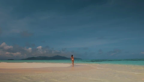 Solitary figure in orange standing in shallow turquoise water on a pristine beach with mountains visible across the water.