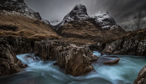 Snow-capped mountains frame a turquoise river flowing over dark rock formations.