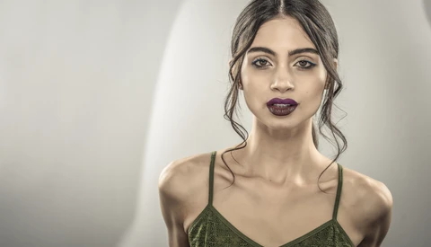 Woman with dark wavy hair and burgundy lipstick wearing olive green camisole against neutral backdrop.