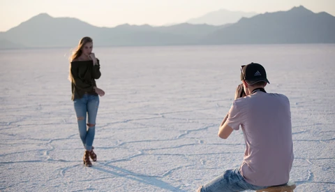 Male photographer seated on a stool captures a female model posing on a salt flat with mountains and water in the background.