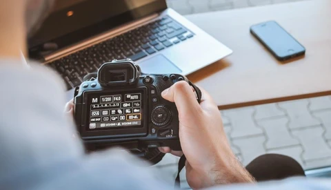 Photographer holding DSLR camera displaying rear LCD screen with menu settings visible.