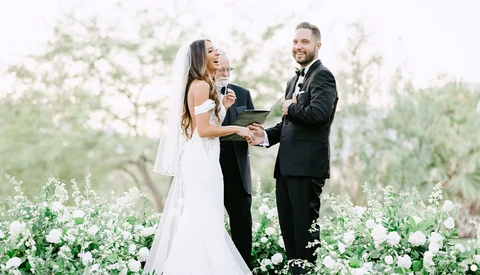 Bride and groom exchange vows during outdoor ceremony surrounded by white flowering plants.