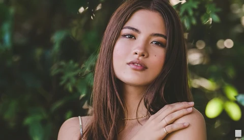 Young woman with long brown hair in natural outdoor lighting surrounded by green foliage.