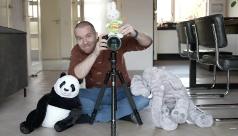 Photographer holding a telephoto lens on a tripod while posing with stuffed animals in an indoor studio setting.