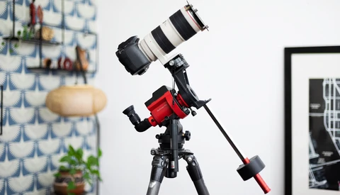 A telescope mounted on a red equatorial mount with a telephoto lens attached, positioned in an indoor studio setting.